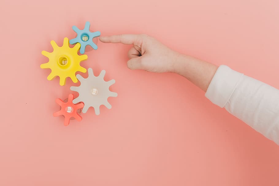 Finger Turning Colorful Toy Gears On Peach Color Wall, - HD Wallpaper 